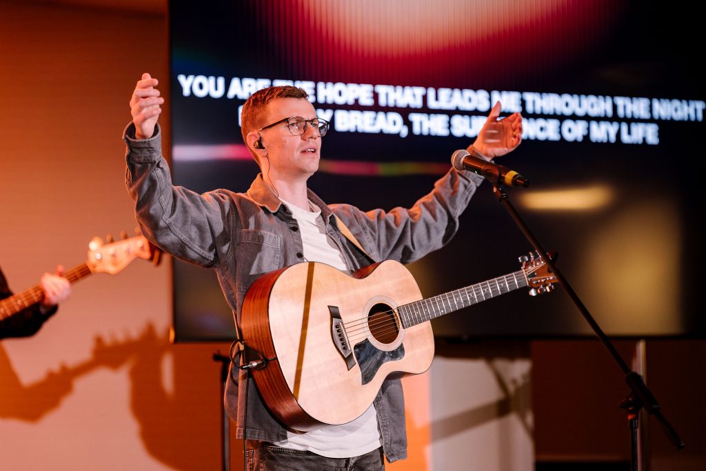 a man with glasses standing on stage with an accoustic guitar, both his hands are raised towards the sky
