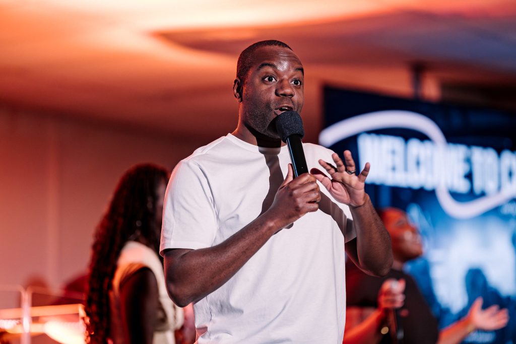 a man standing on stage speaking into a microphone, the words "welcome to church" can be seen on a screen behind him