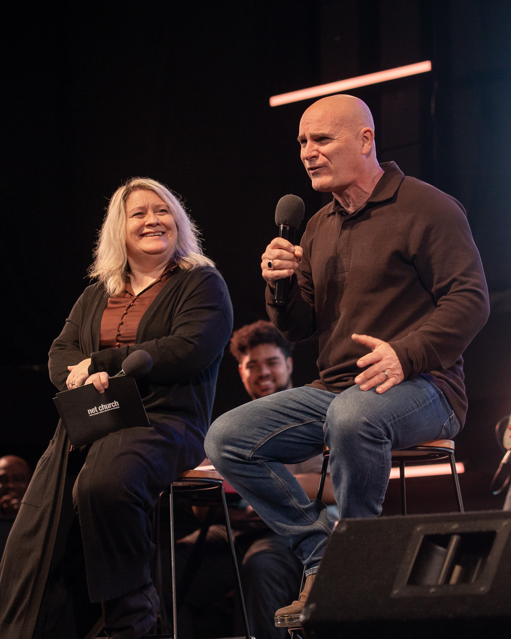 two speaker sitting on stools on stage. the one on the left is a woman looking towards a man on the right, both are smiling.